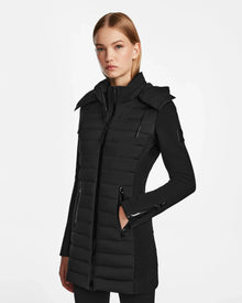 A woman with straight blonde hair models the RUDSAK MERINA - 8122546 BLACK, a long, fitted quilted down jacket with a high collar and zippered pockets, standing against a white background.