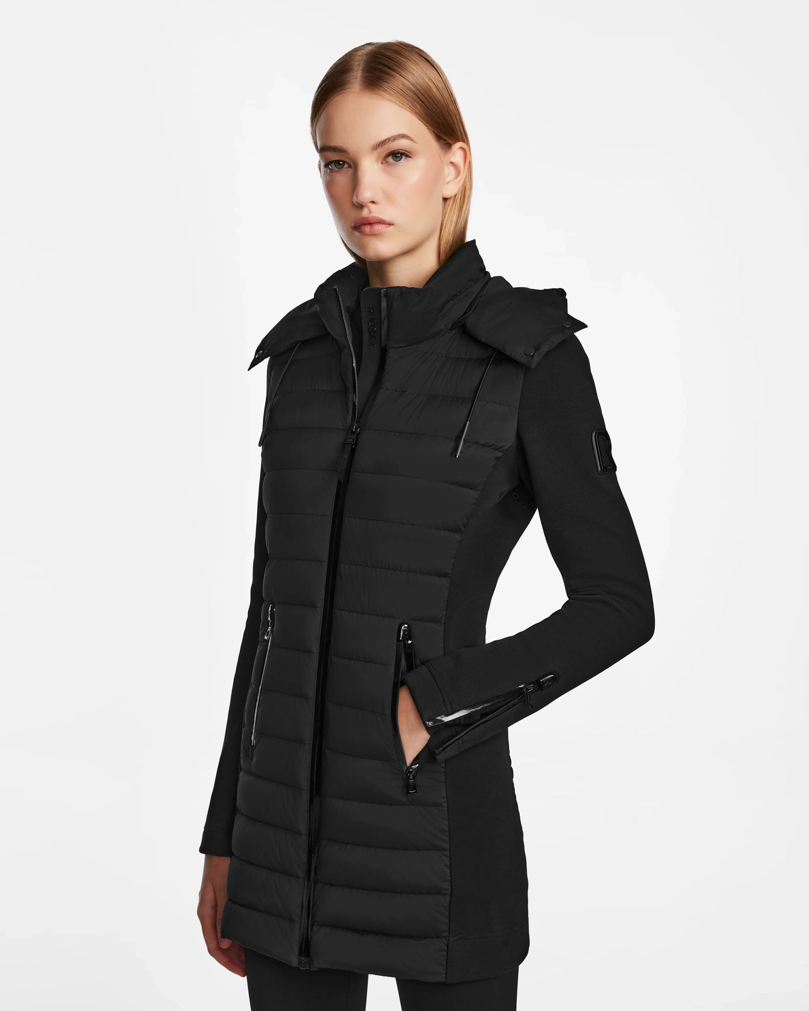 A woman with straight blonde hair models the RUDSAK MERINA - 8122546 BLACK, a long, fitted quilted down jacket with a high collar and zippered pockets, standing against a white background.