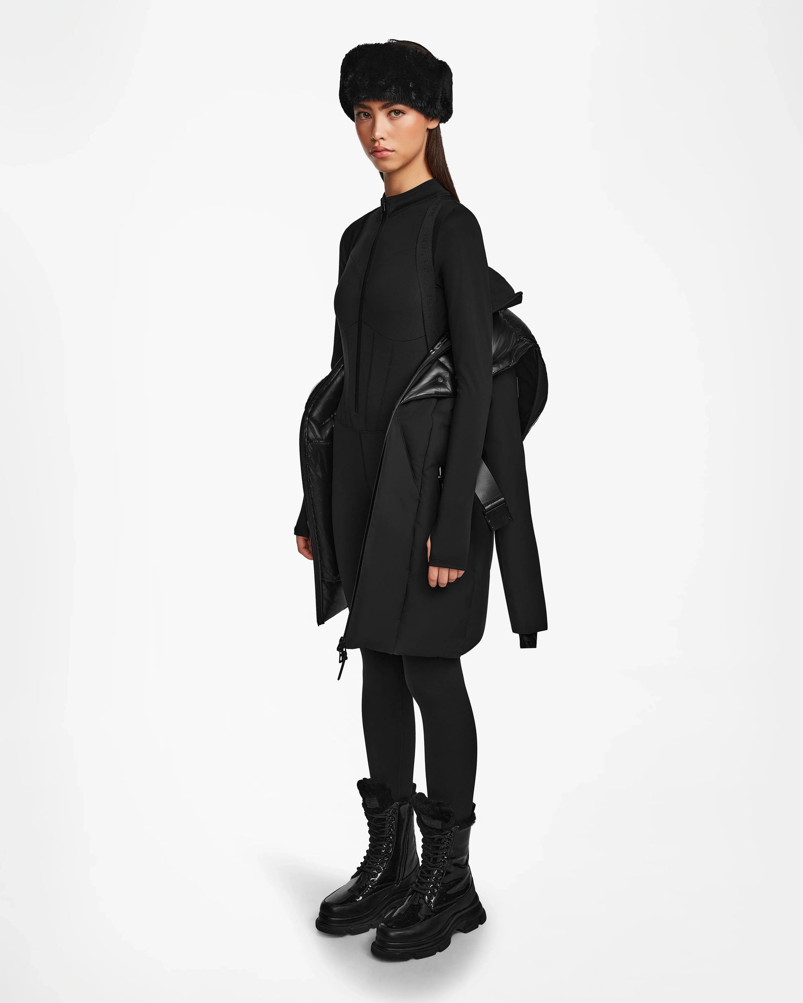 Wearing the Rudsak MODA M - 8124555 Black down parka draped over one shoulder, a person stands against a plain white background, styled with a black fur hat, long-sleeve dress, black leggings, and chunky boots.