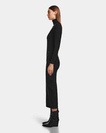 A woman stands in profile against a plain background, wearing a fitted black turtleneck, an elegant MONICA - 8524516 Black skirt by Rudsak, and black buckle ankle boots.