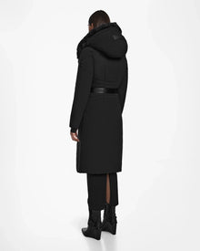A person faces away, wearing the Rudsak MUSA - L124534 Black women’s long puffer coat with RDS-certified down, fur-trimmed hood, belted waist, black pants, and boots against a plain white background.
