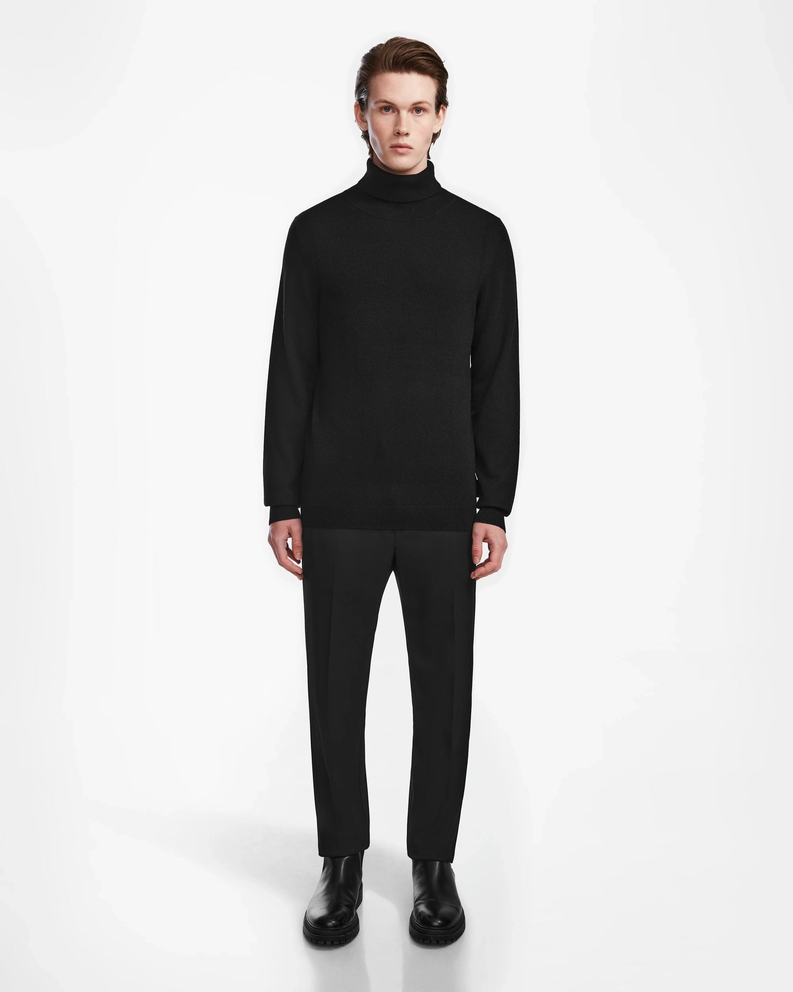 A person stands facing forward against a plain white background, wearing the Rudsak NICOLAS - 6524528 Black mens cashmere turtleneck sweater, paired with black pants and black boots.