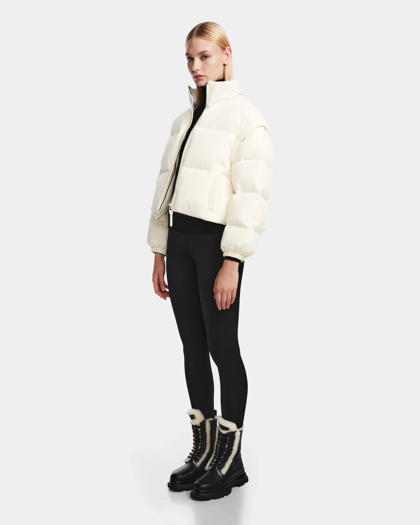 A woman wears the Rudsak OPRAH - 8124708 Iceland cropped down puffer, styled with black leggings and black winter boots with white fur trim, standing against a plain light gray background.