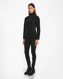 A woman with straight brown hair and a neutral expression stands against a white background, wearing the Rudsak ORCHID - 8524616 Black women’s half-zip pullover with vegan leather trims, black leggings, and black ankle boots.