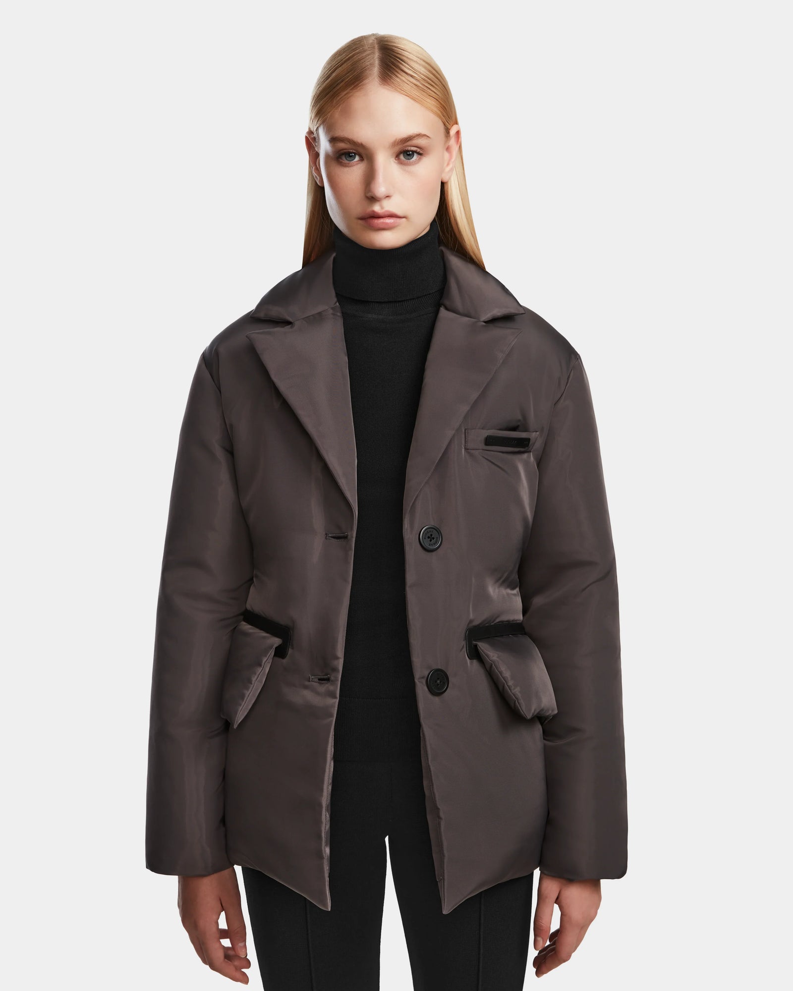 A woman with straight blonde hair wears the Rudsak PAOLA - 8124547 Grey oversized womens down blazer-style puffer jacket over a black turtleneck and black pants, standing against a plain white background.