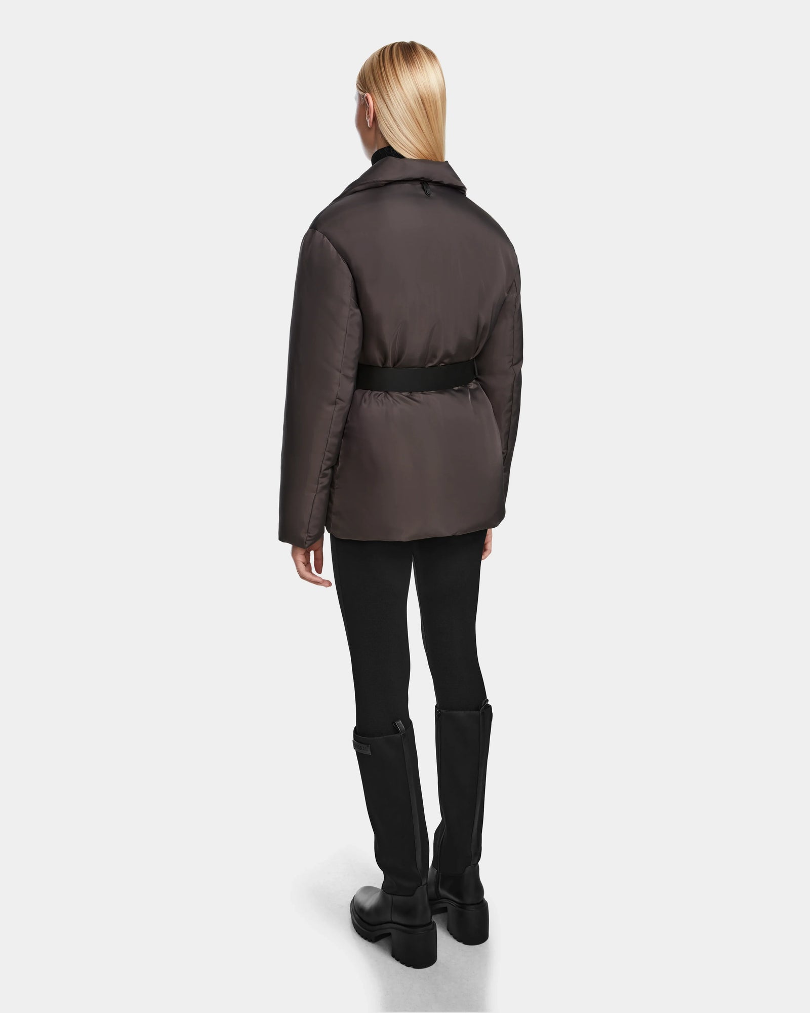 A person with straight blonde hair, dressed in the Rudsak PAOLA - 8124547 Grey RDS-certified down puffer jacket, black leggings, and black knee-high boots stands facing away against a plain light background.