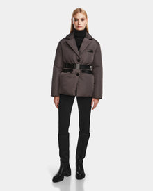 A woman with straight blonde hair wears the Rudsak PAOLA - 8124547 Grey down blazer over a black turtleneck, slim black pants, and knee-high boots, standing against a plain white background.