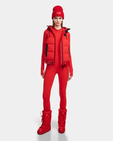 A woman stands facing forward in a Rudsak RAQUEL M - 8124538 Flame Red outfit, featuring a reversible ski vest, beanie, fitted jumpsuit, and puffy boots against a plain white background.