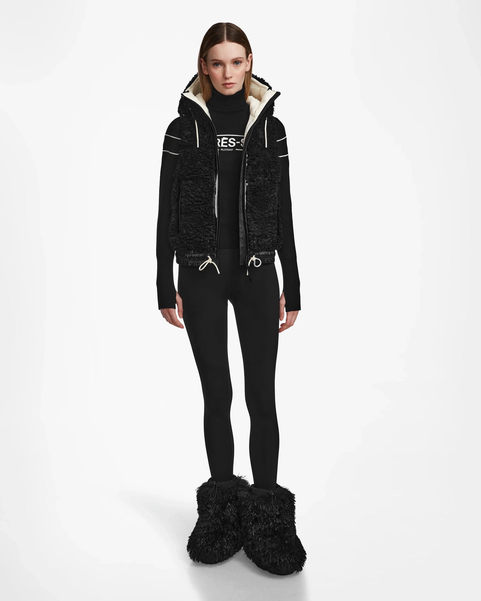 A woman faces forward in the Rudsak RAQUEL S - 8124526 Black vest with a black long-sleeve, black leggings, and oversized black furry boots—an ideal winter mountain look—all against a plain white background.