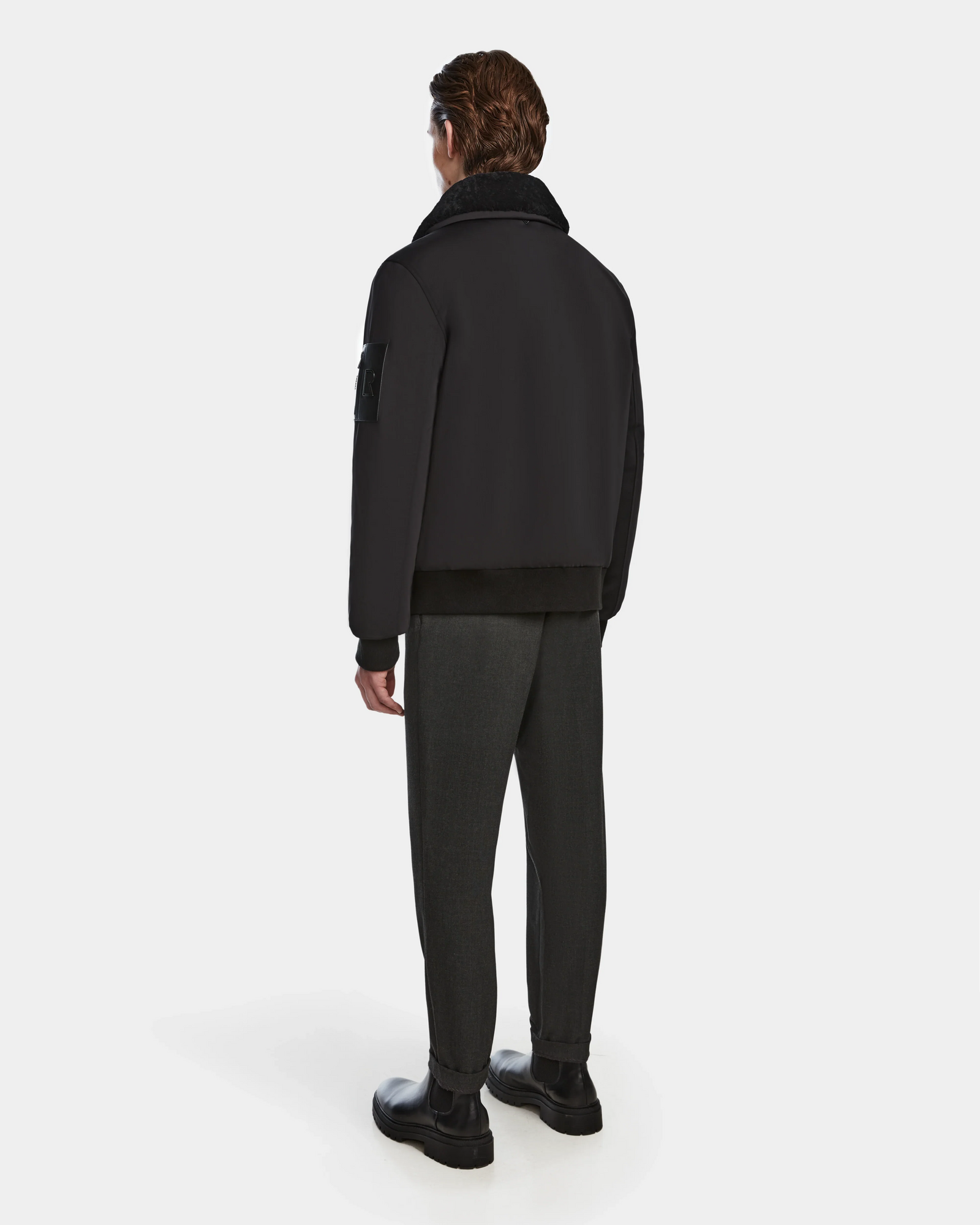 A man stands facing away in a Rudsak REECE - 6124592 Black mens bomber jacket with a shearling collar, paired with gray pants and black boots, set against a plain white background.