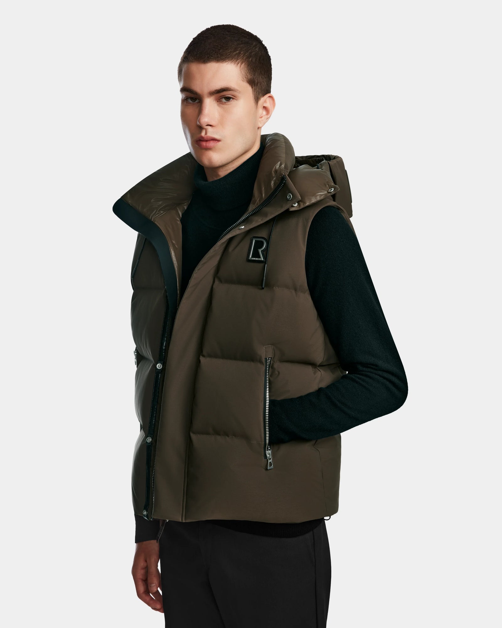 A young man stands facing forward with a neutral expression, wearing the Rudsak REN M - 9123512 Army M weather-resistant dark olive green puffer vest over a black turtleneck against a plain light background.