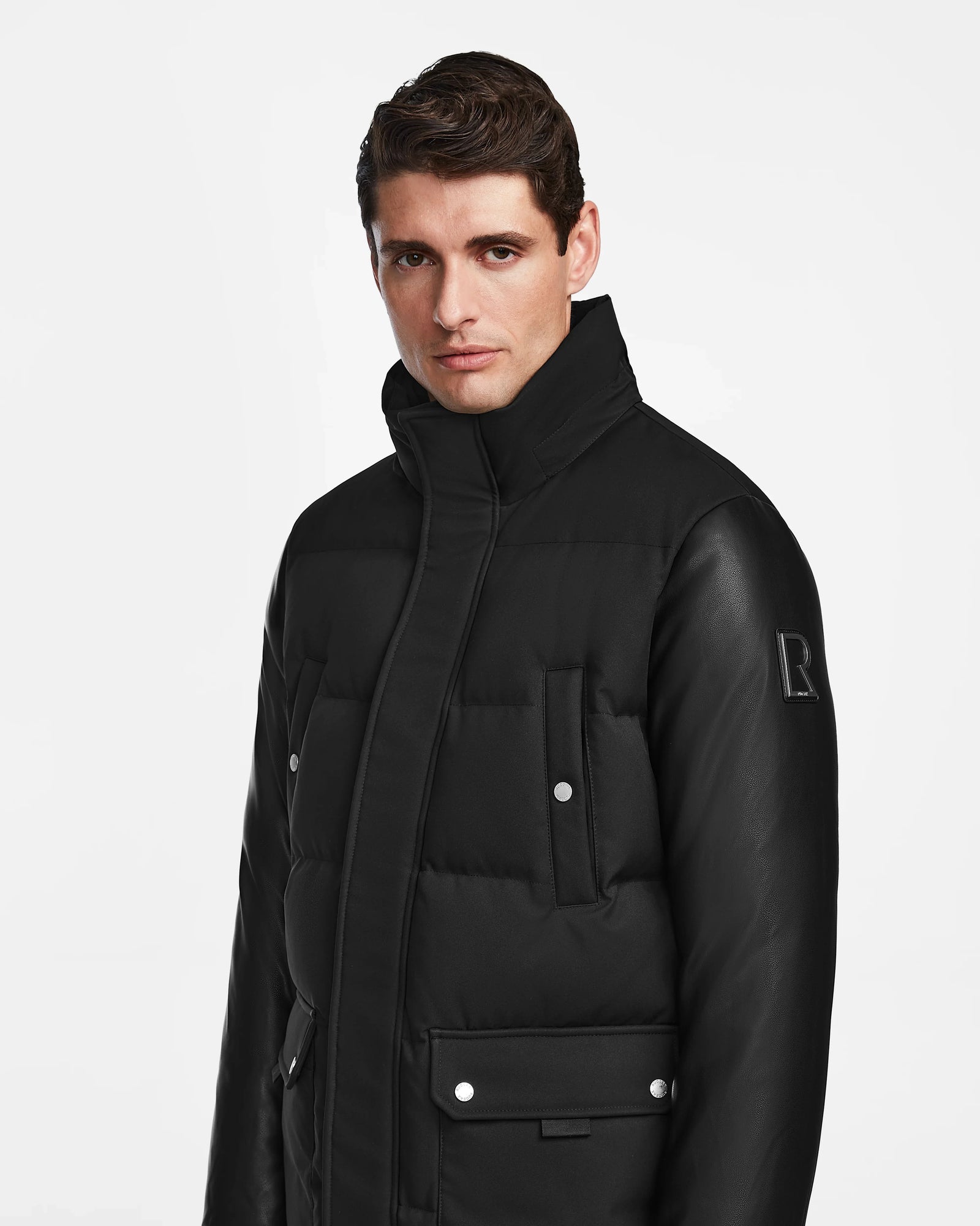 A man with short brown hair wears the Rudsak Ricky R - M123506 Black men's quilted down parka with recycled leather sleeves, a high collar, buttoned pockets, and a subtle R logo on the sleeve, standing against a plain white background.