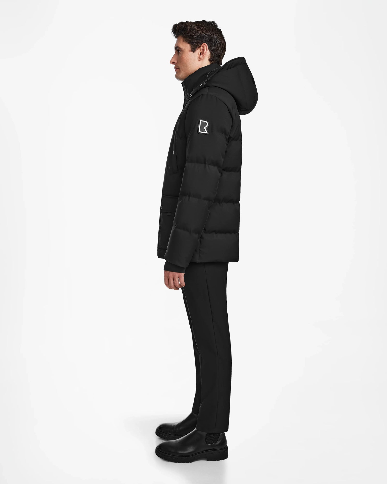 A person stands in profile against a white background wearing the Rudsak RICKY - M123502 Black parka, black pants, and black boots. The winter parka features a white R logo on the upper sleeve.