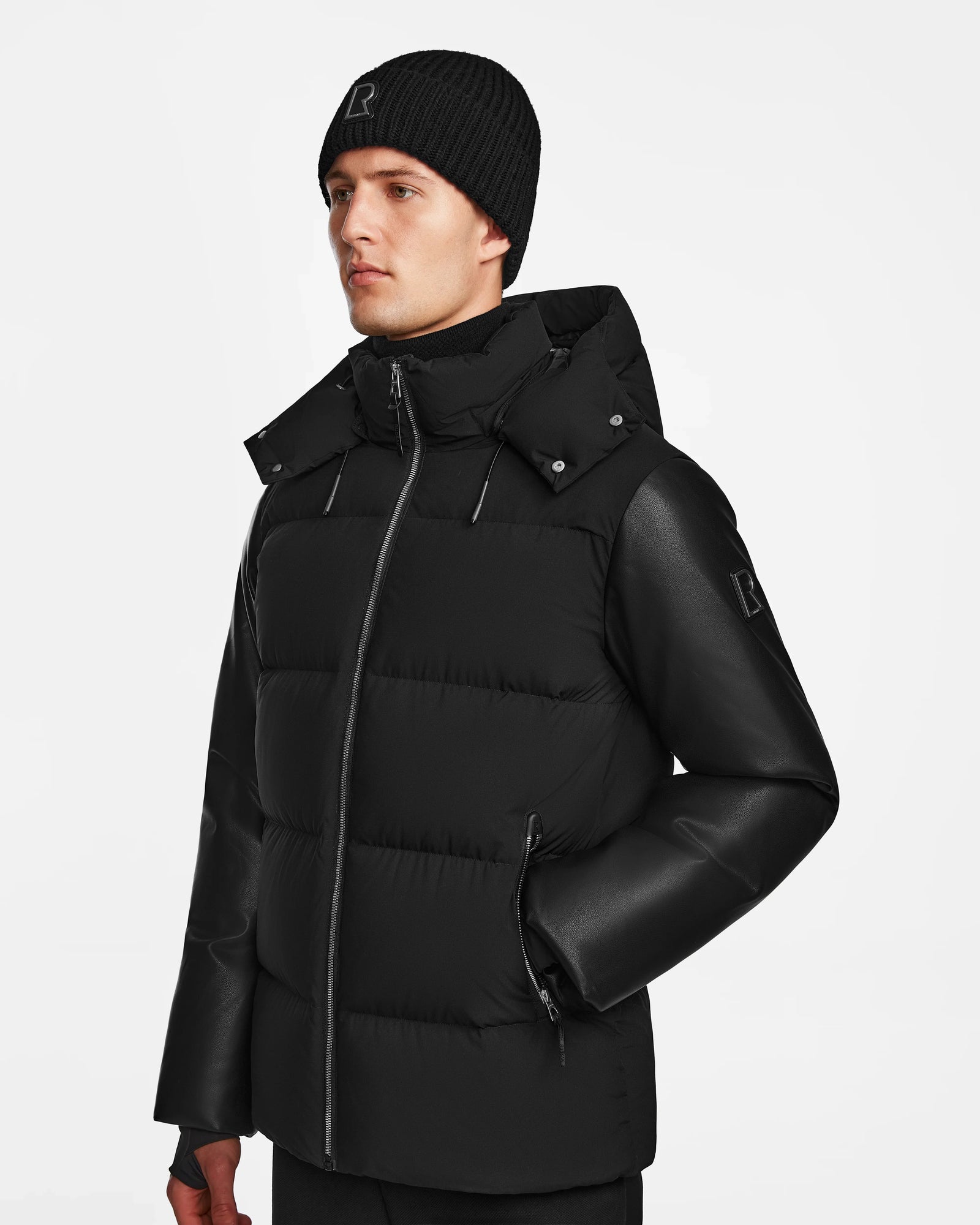 A man wearing the Rudsak RONNY M - M124513 Black puffer jacket with a hood and a black knit beanie stands against a plain white background, facing slightly left with a neutral expression.