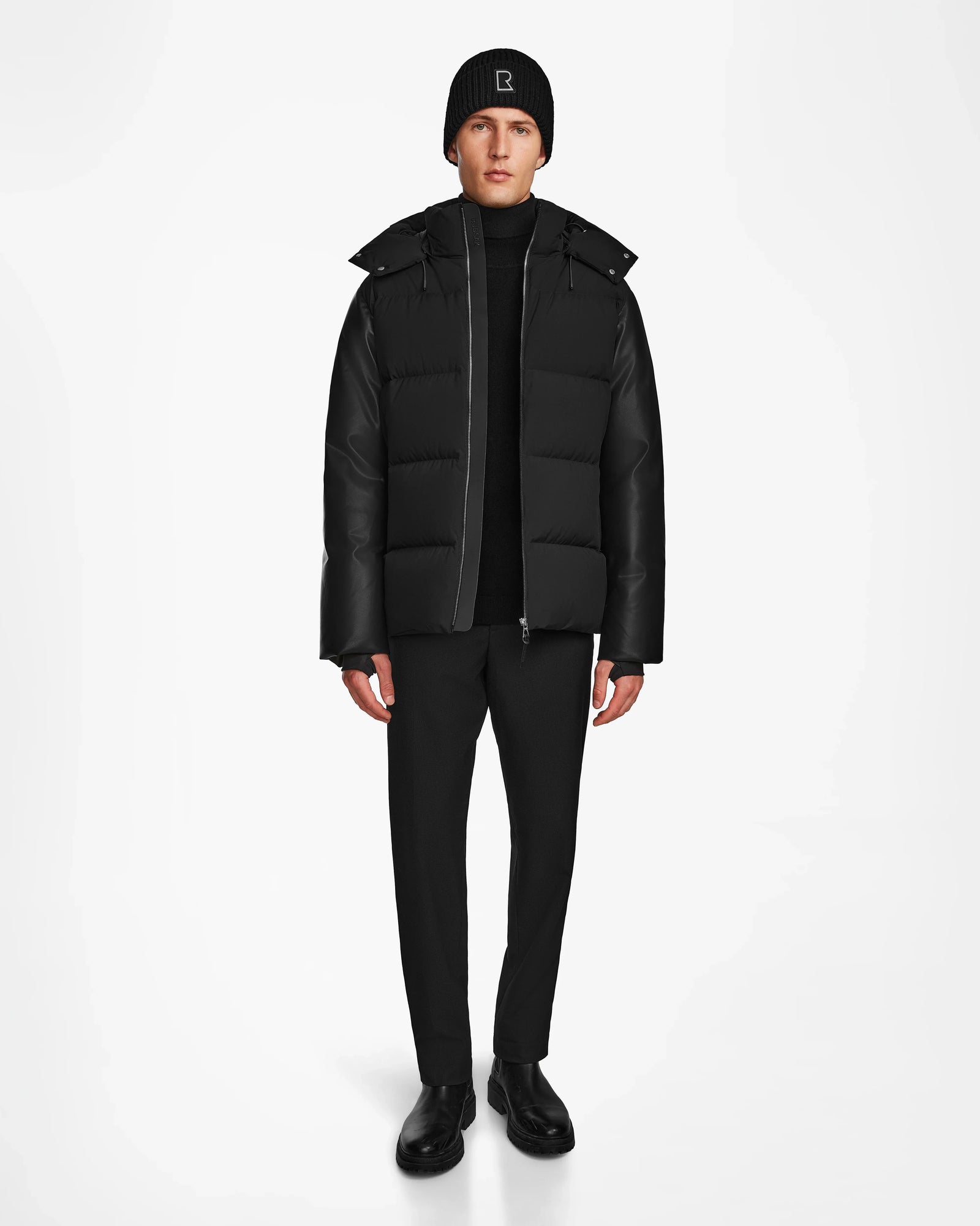 A person wears the Rudsak RONNY M - M124513 Black puffer jacket, paired with black pants, boots, and a beanie, standing against a plain white background facing forward with arms at their sides.