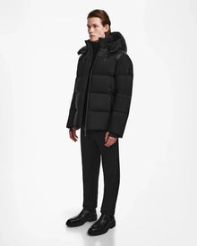 A person stands against a white background wearing the Rudsak THEO - M125531 Black men's down puffer with high collar and hood, paired with black pants and shoes—a sleek, minimalist, weather-resistant outfit for any season.