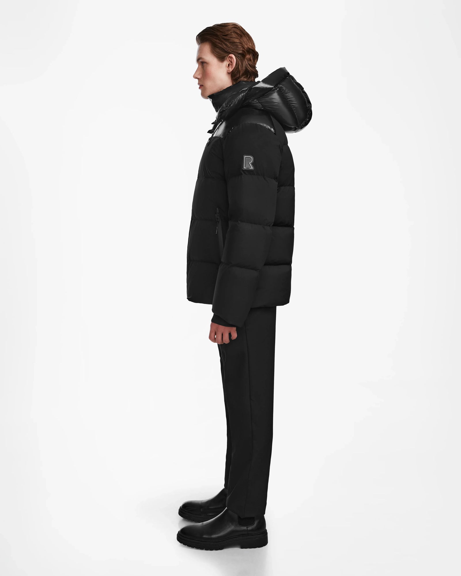 A person stands in profile wearing the Rudsak THEO - M125531 Black weather-resistant puffer jacket with a hood, paired with black pants and black shoes against a plain white background.