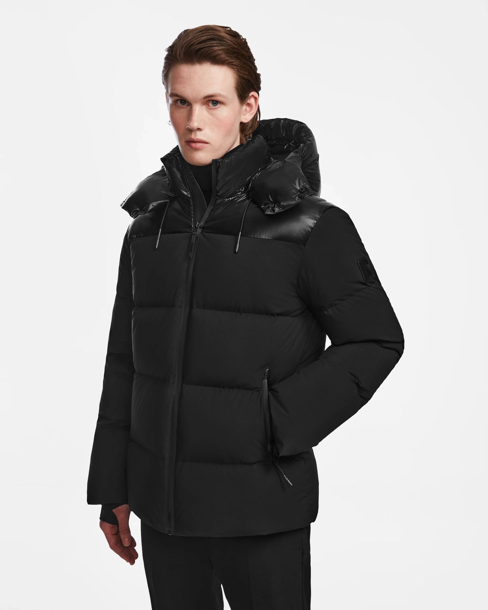 A light-skinned person with straight brown hair models the Rudsak THEO - M125531 Black men's down puffer hooded jacket, standing against a plain white background and gazing slightly to the side.