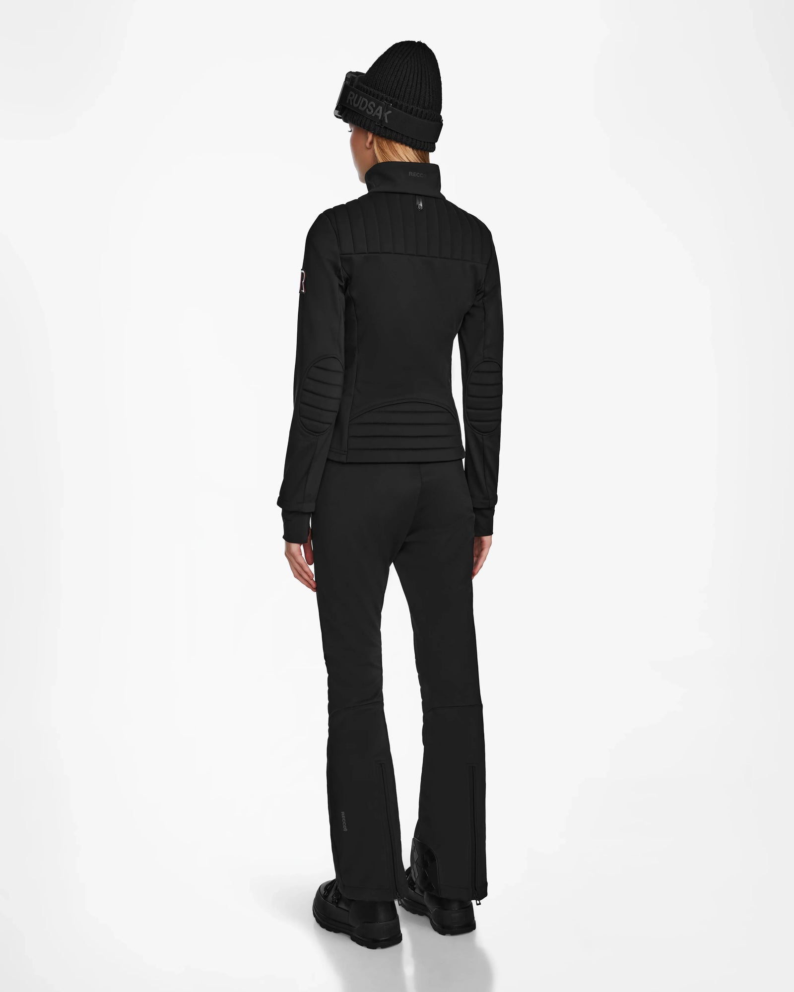 A person stands facing away against a plain white background, wearing the Rudsak ROSALIE - 8124784 Black ski jacket as part of an all-black winter outfit with matching beanie and pants.