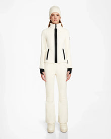 A woman faces forward in the ROSALIE - 8124784 Iceland women's fitted ski jacket by Rudsak with black accents, matching pants, a knit beanie, and platform boots. The high-performance jacket features RECCO rescue technology for extra safety.