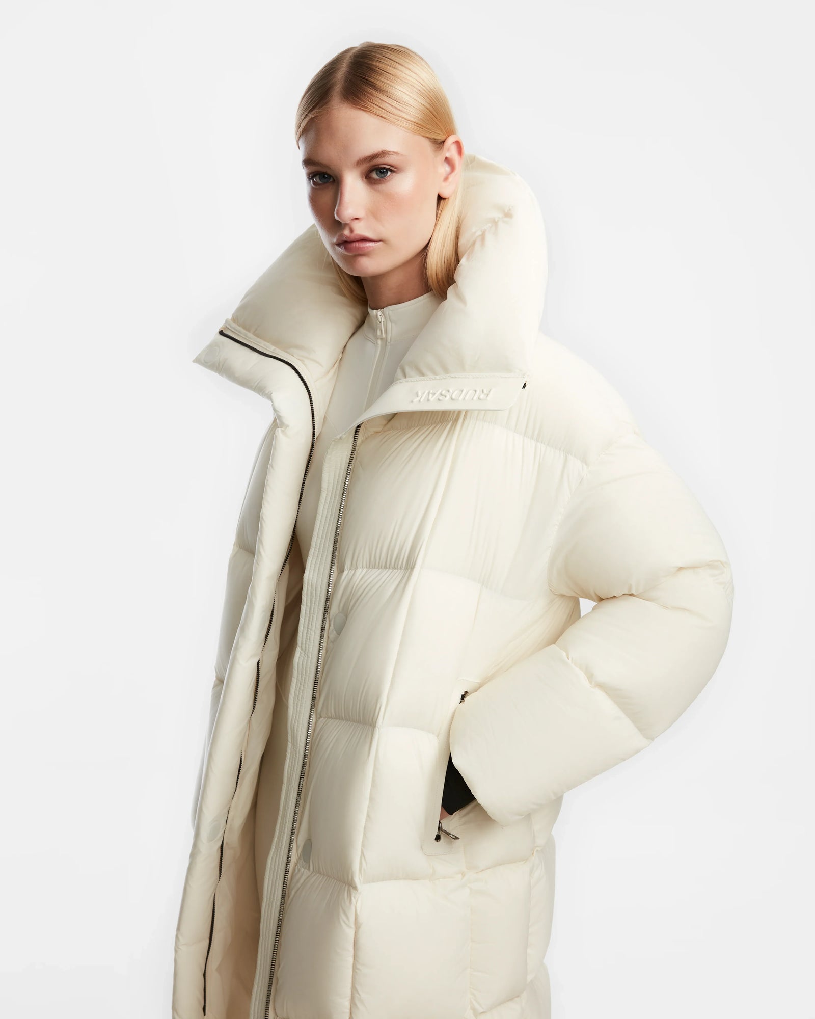 A woman with straight blonde hair wears the Rudsak SASH - 8124536 Iceland long, cream-colored womens down puffer coat with a high collar, hands in the pockets, standing against a plain white background and displaying a neutral expression.