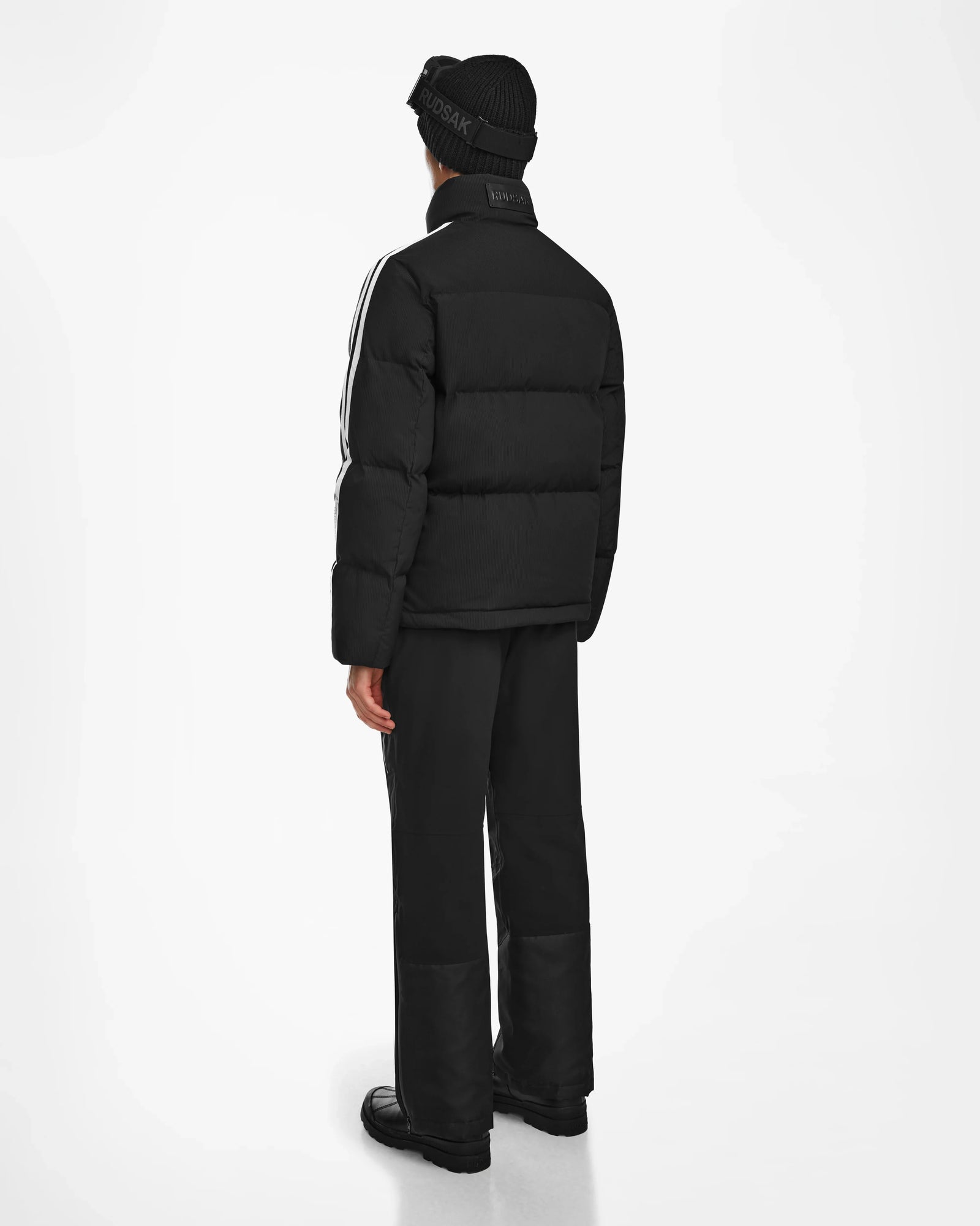 A person faces away, wearing the Rudsak USHER - 6124516 Black RDS-certified down jacket with white sleeve stripes, black pants, shoes, and beanie hat. The background is plain white.