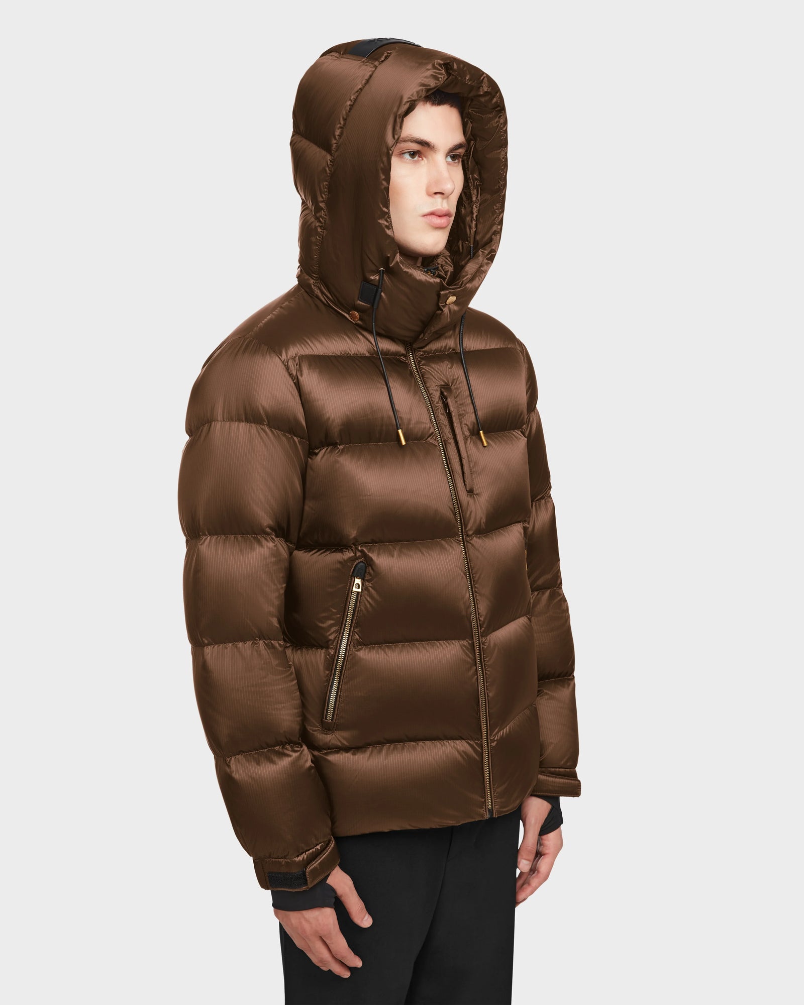 A young man wearing the Rudsak VINCE S - 6124560 Antique Bronze men’s quilted down puffer with a shiny finish, hood, and zippered pockets stands against a light background, facing slightly to the side.