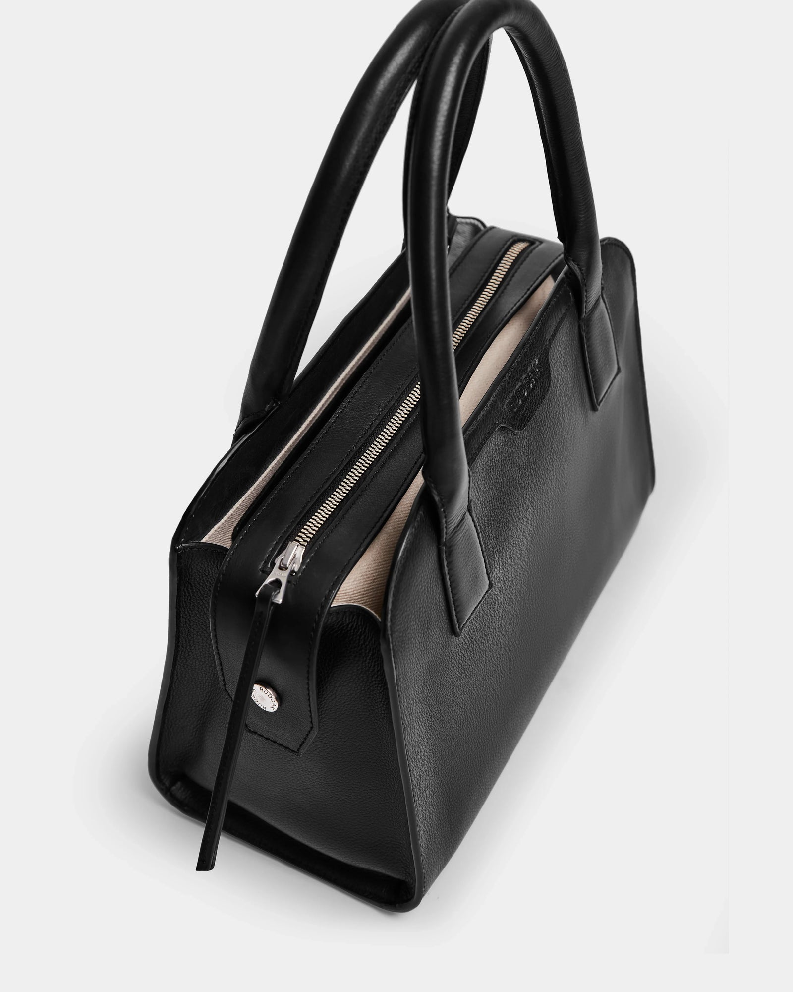 The YASMINA - 8324527 Black by Rudsak is a womens leather tote bag with sturdy double handles and a silver zipper, shown from above on a white background. Its interior is lined with light-colored fabric.