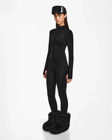 A woman poses against a white background in the Rudsak ZADIE - 8524609 Black ski catsuit, black ski goggles atop her head, and oversized black furry boots, with her arms resting by her sides.