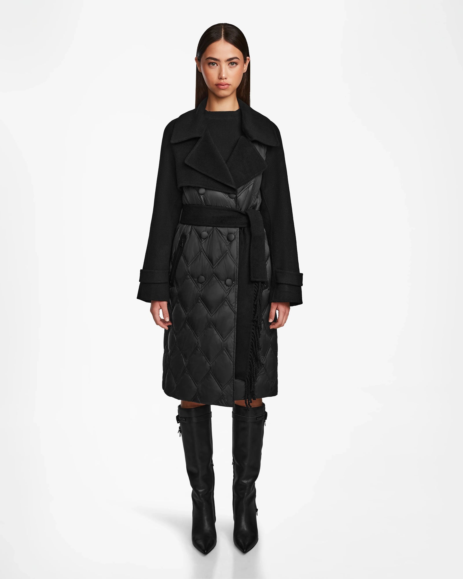 A woman faces forward in the Rudsak ZOE - 8124711 Black quilted down trench coat with a belted waist and tall black boots, set against a plain white background.