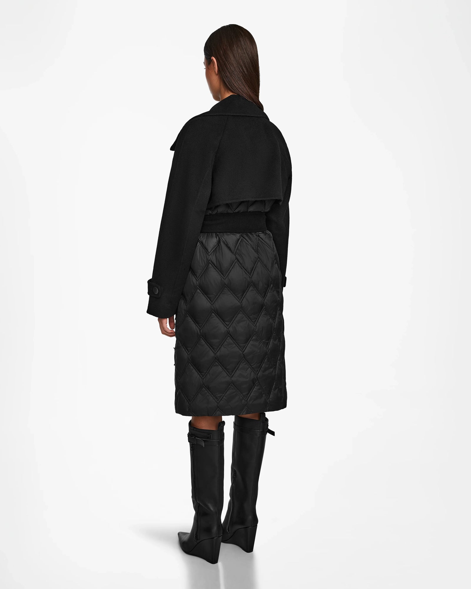 A woman faces away wearing the Rudsak ZOE - 8124711 Black womens trench coat, featuring a quilted lower half, paired with tall black boots against a plain white background.