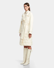 A woman stands in profile wearing the Rudsak ZOE - 8124711 Iceland cream trench coat with quilted detailing, paired with matching knee-high boots against a plain light gray background.