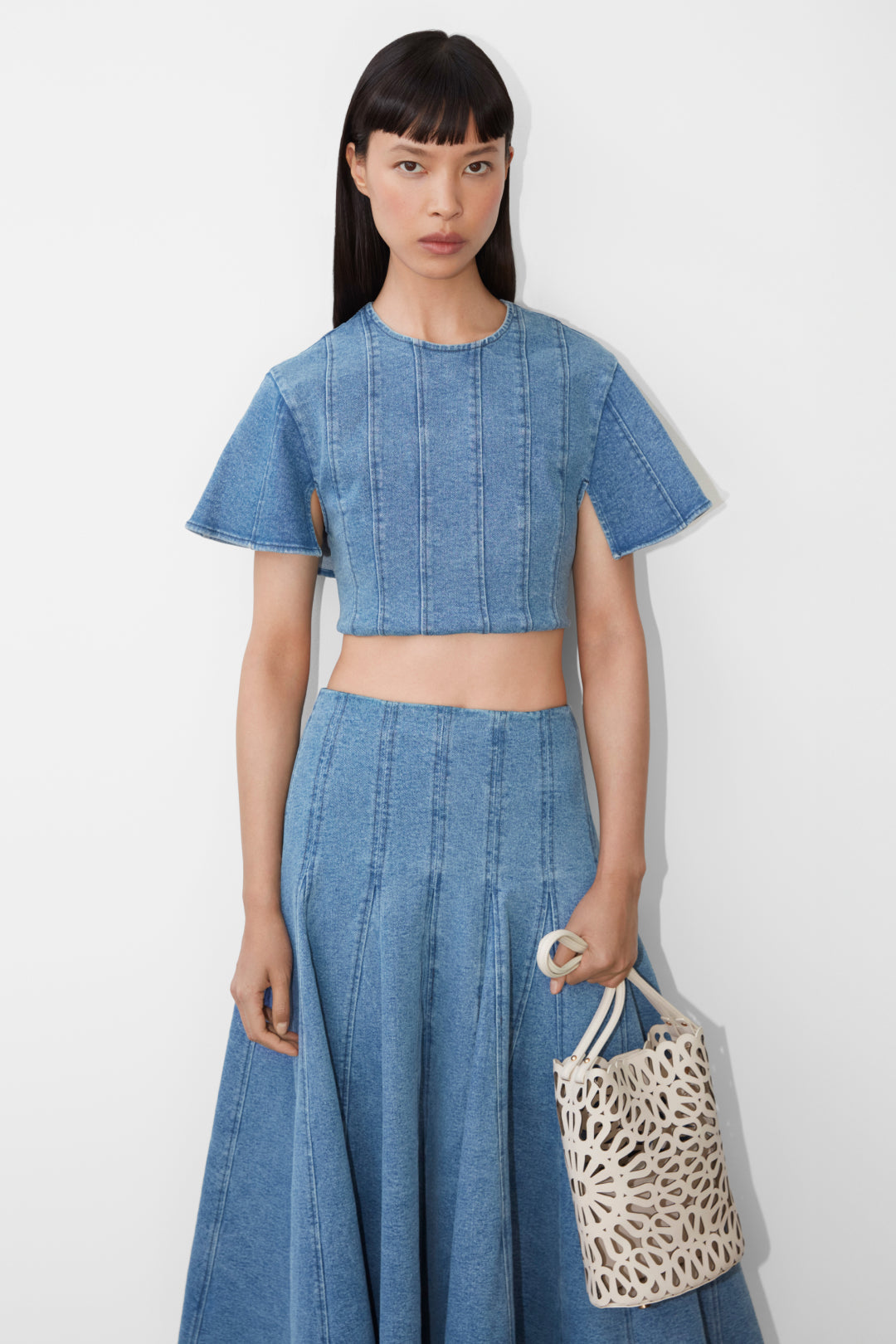 Woman wearing a light blue denim crop top with vertical seams and short sleeves paired with a matching pleated denim skirt.