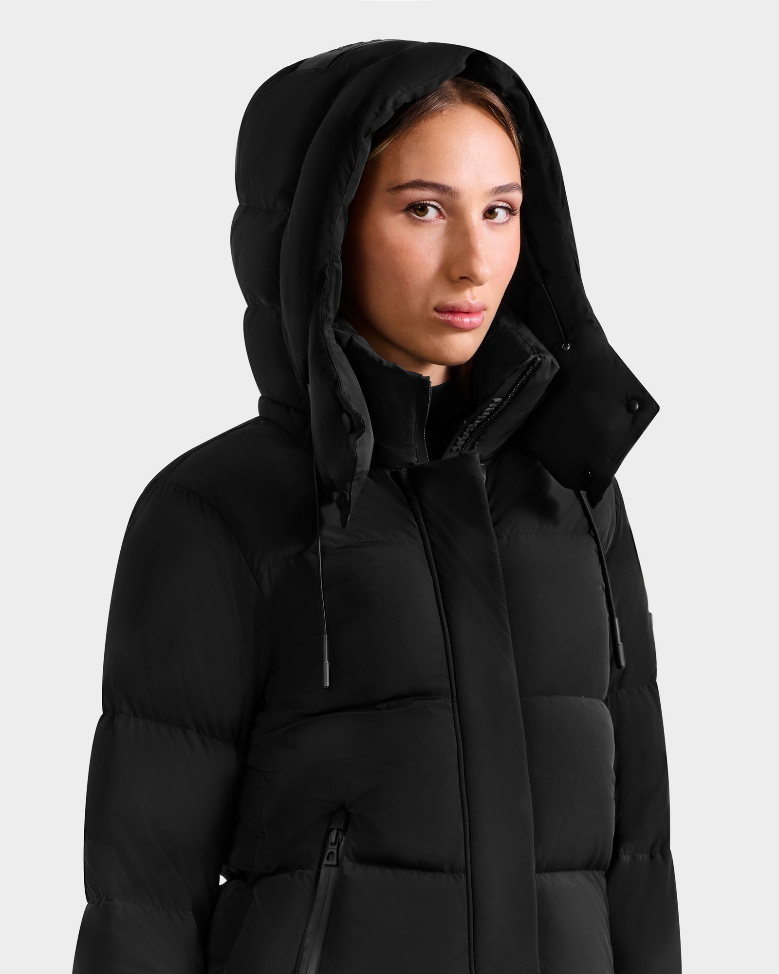 A young woman with light skin and brown hair models the Rudsak ALESSANDRA S-M - L125551 Black down puffer jacket with the hood up, gazing neutrally at the camera against a plain light gray background—a true cold-weather essential.