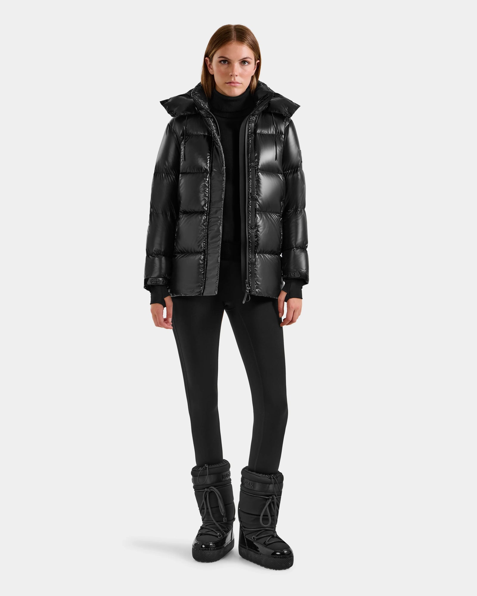 A woman faces forward against a light gray background, wearing the Rudsak ALESSANDRA S-S - L125560 Black glossy puffer jacket, paired with black pants, a black turtleneck, and black winter boots.