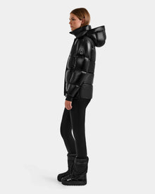 A woman wearing the Rudsak ALESSANDRA S-S - L125560 Black glossy puffer jacket, black leggings, and black winter boots stands in profile against a plain white background.