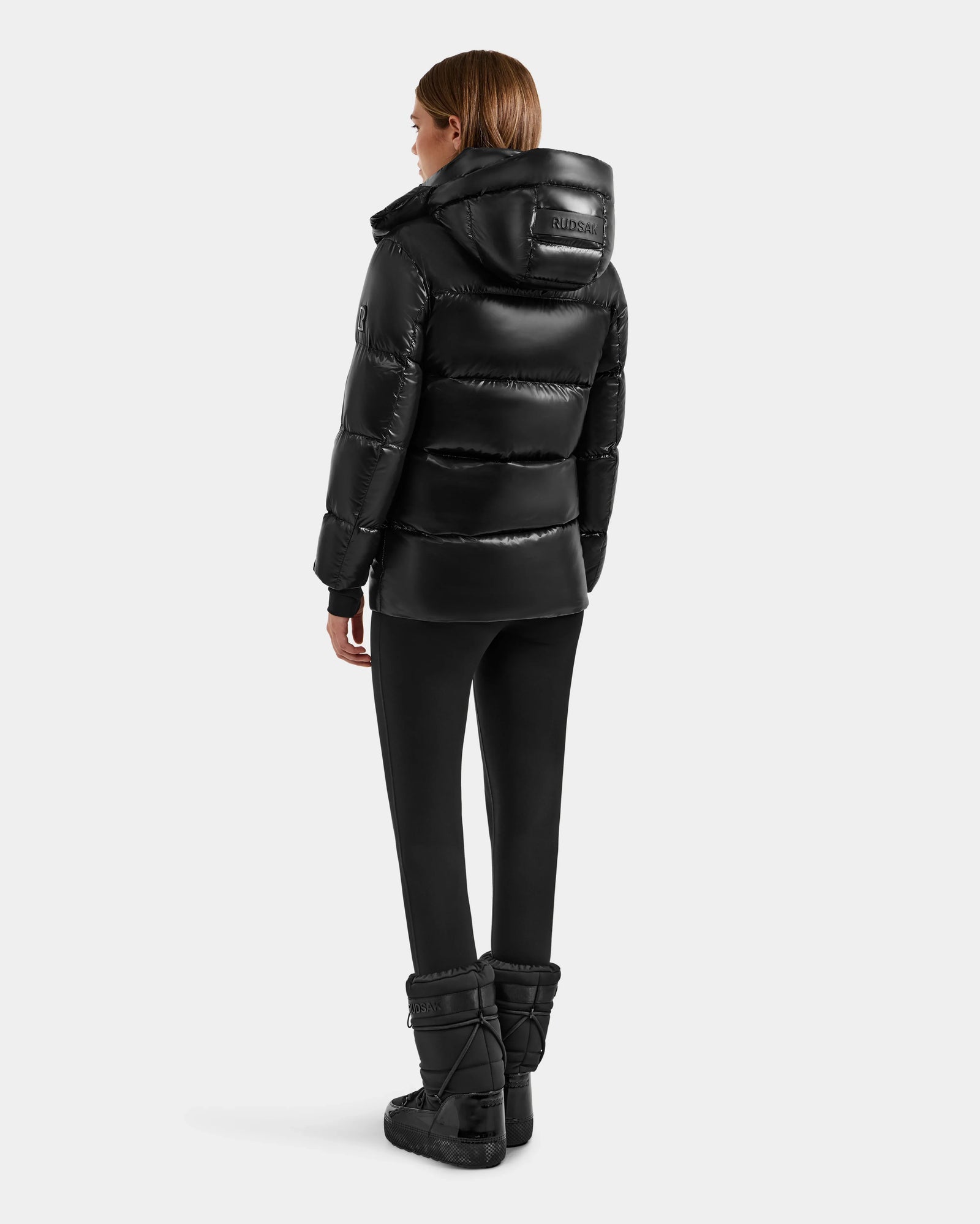 A person wears the Rudsak ALESSANDRA S-S - L125560 Black down puffer with hood and back logo patch, paired with black pants and black quilted boots, standing turned slightly to the side.