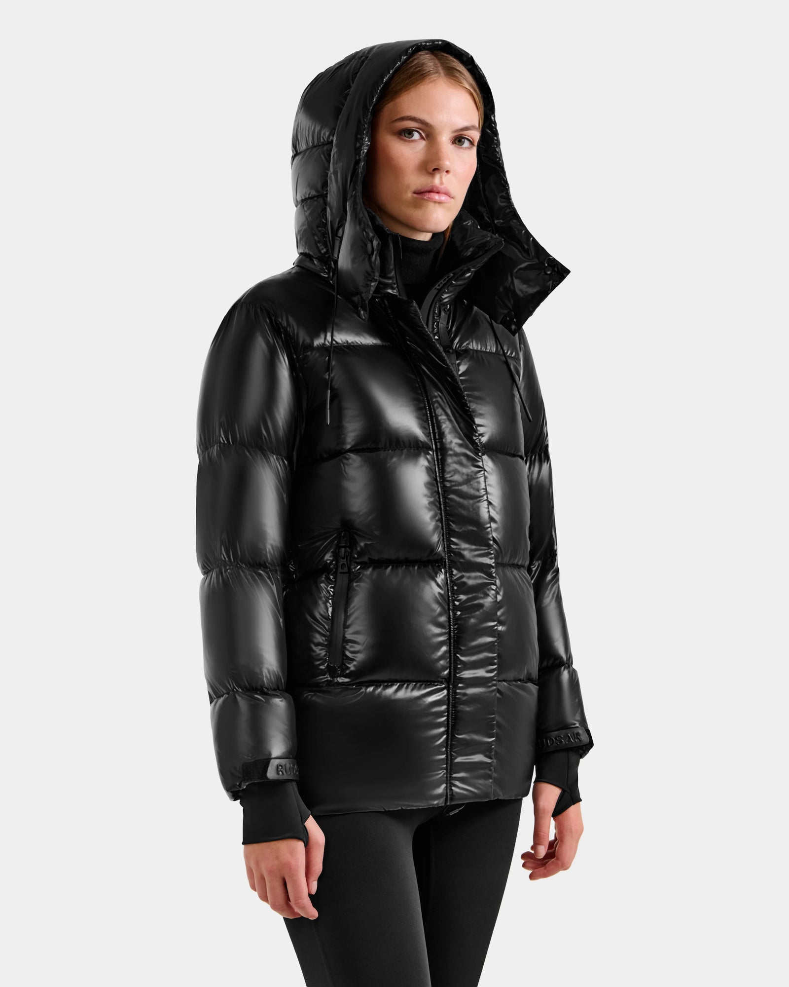 A woman wearing the Rudsak ALESSANDRA S-S - L125560 Black down puffer with a hood stands against a light gray background, gazing slightly to the side with a neutral expression.