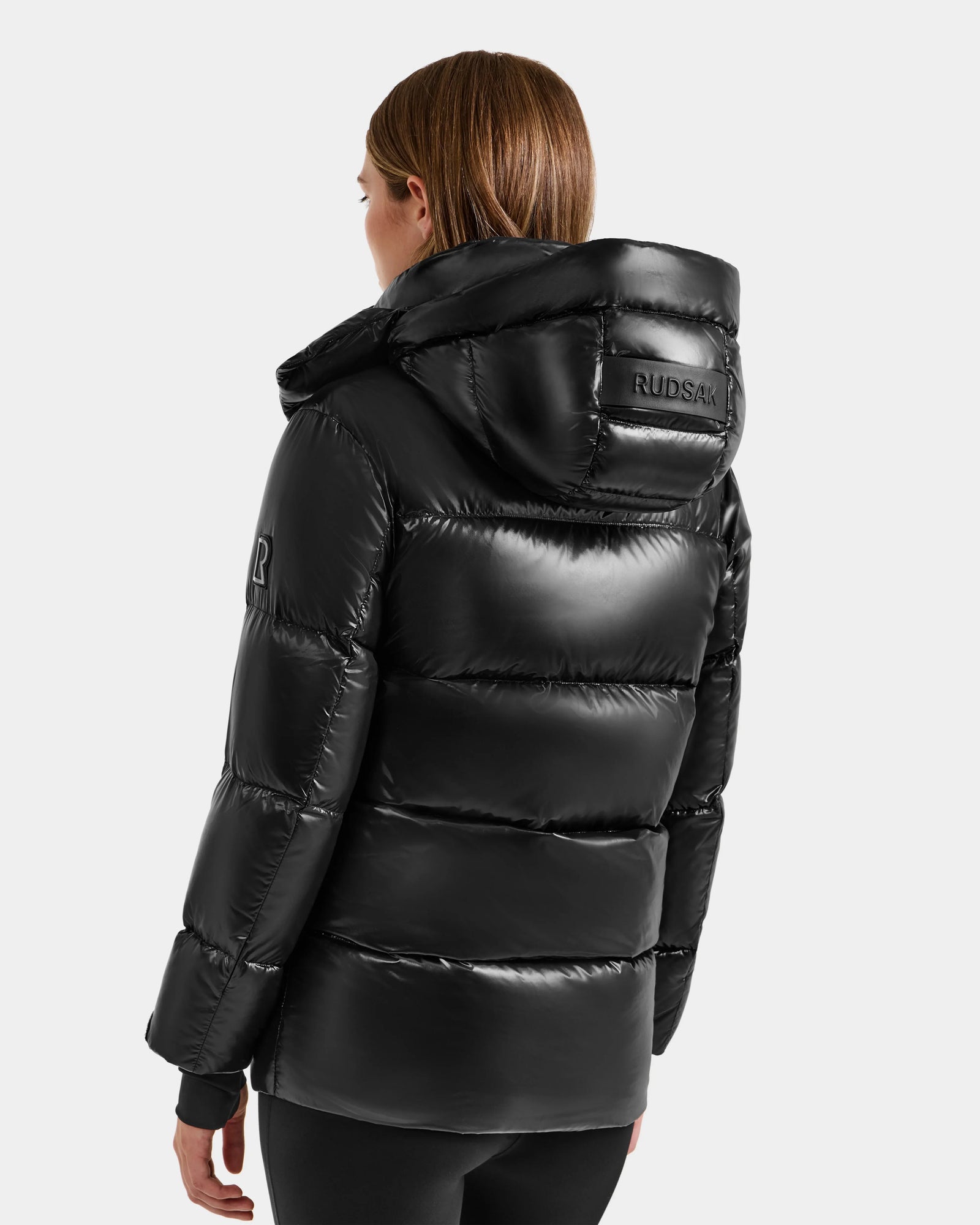 A person with straight, light brown hair wears the Rudsak ALESSANDRA S-S - L125560 Black glossy puffer jacket with hood. The Rudsak logo is visible on the back and left sleeve. The background is plain and light-colored.