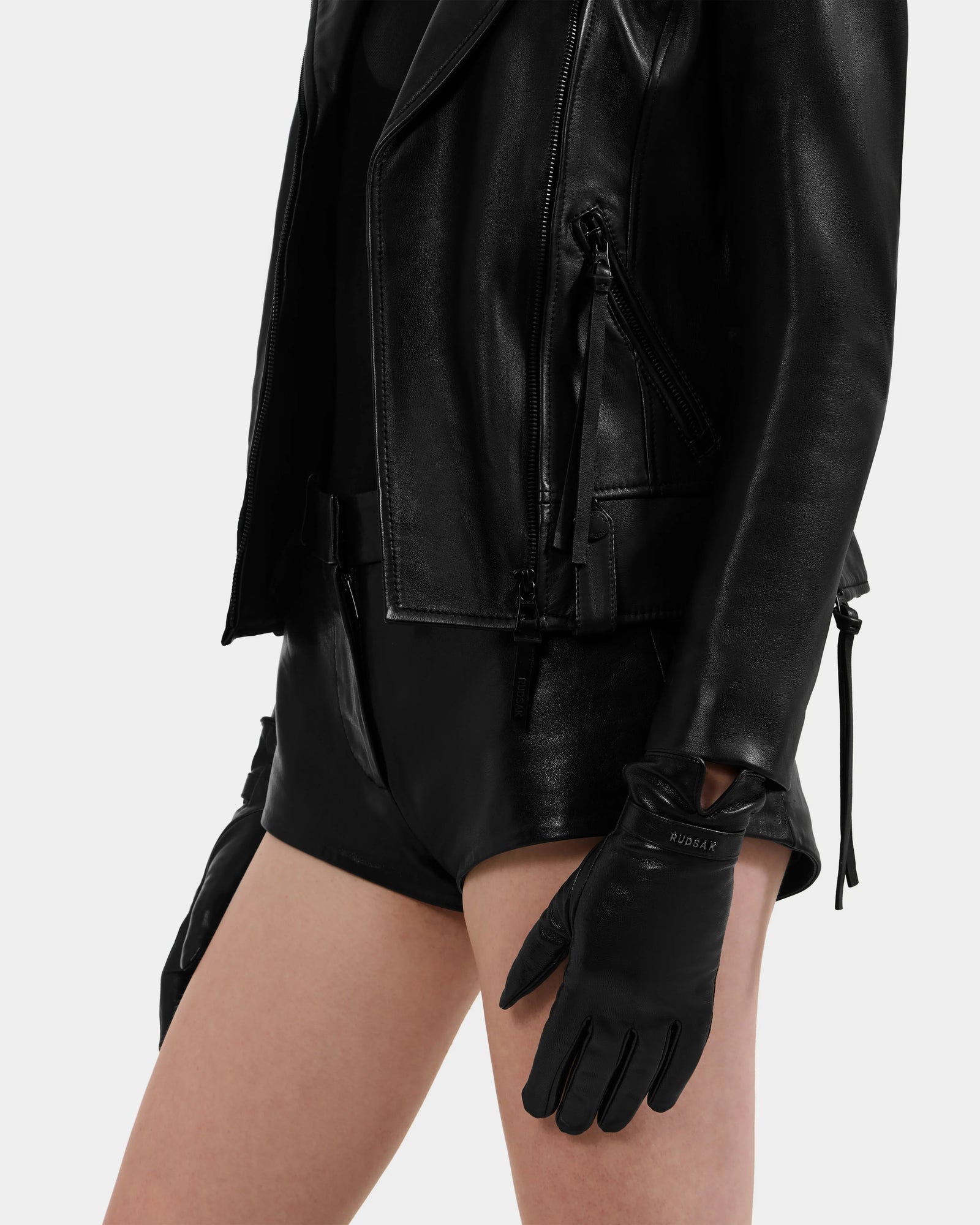 A person stands against a light background, wearing Rudsak’s ALEXIA - L425543 Black leather jacket and sleek women’s gloves, styled with black shorts. Only the torso and upper legs are visible.