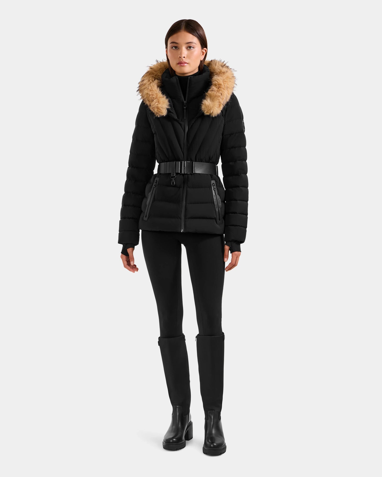 A woman faces forward wearing the Rudsak ALISA F - L125527 Black quilted puffer jacket with a faux-fur trim hood, paired with black leggings and boots, set against a plain light gray background.