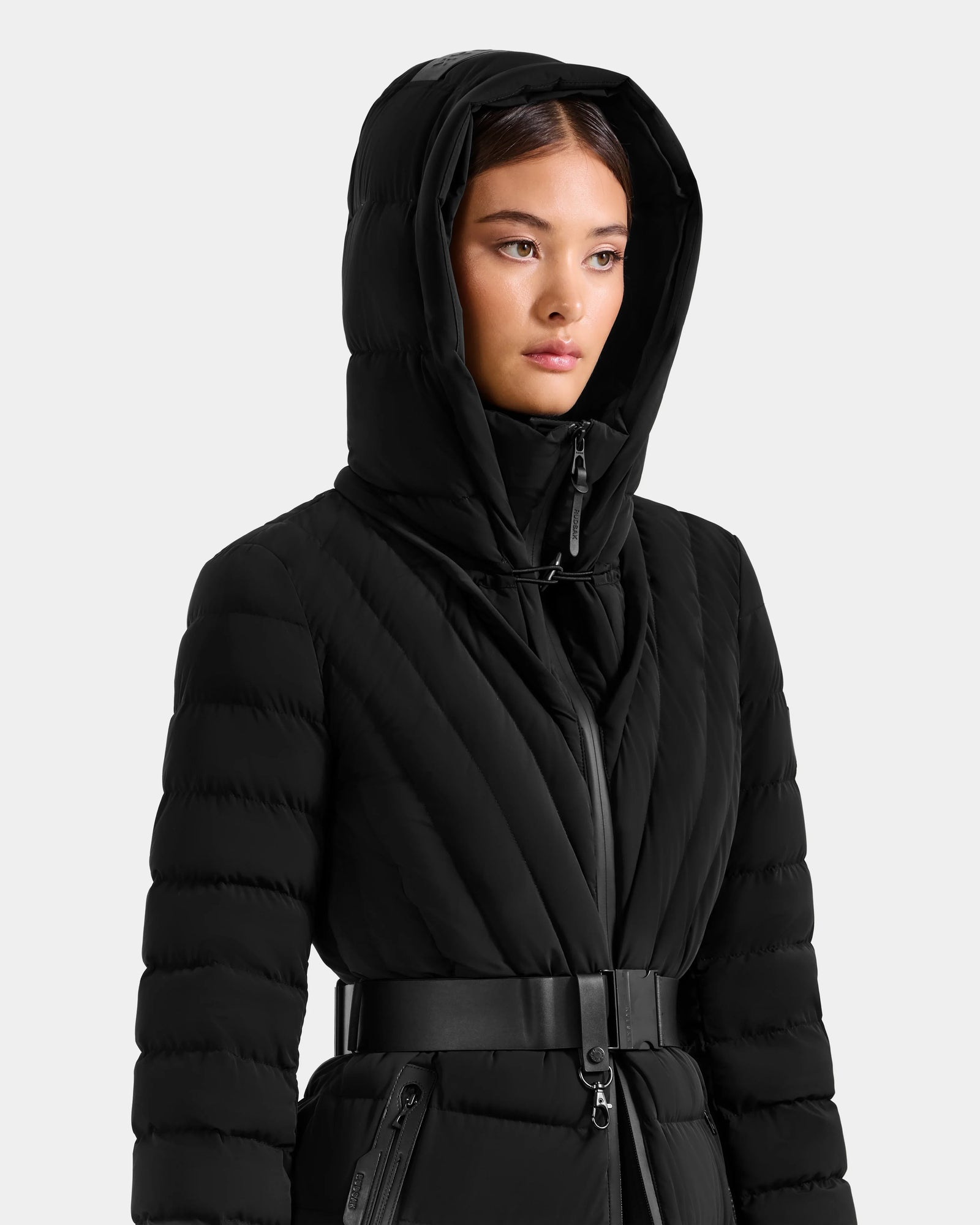 A woman wears the Rudsak ALISA L - L125574 Black quilted puffer coat with a hood, looking slightly to the side against a plain light background.