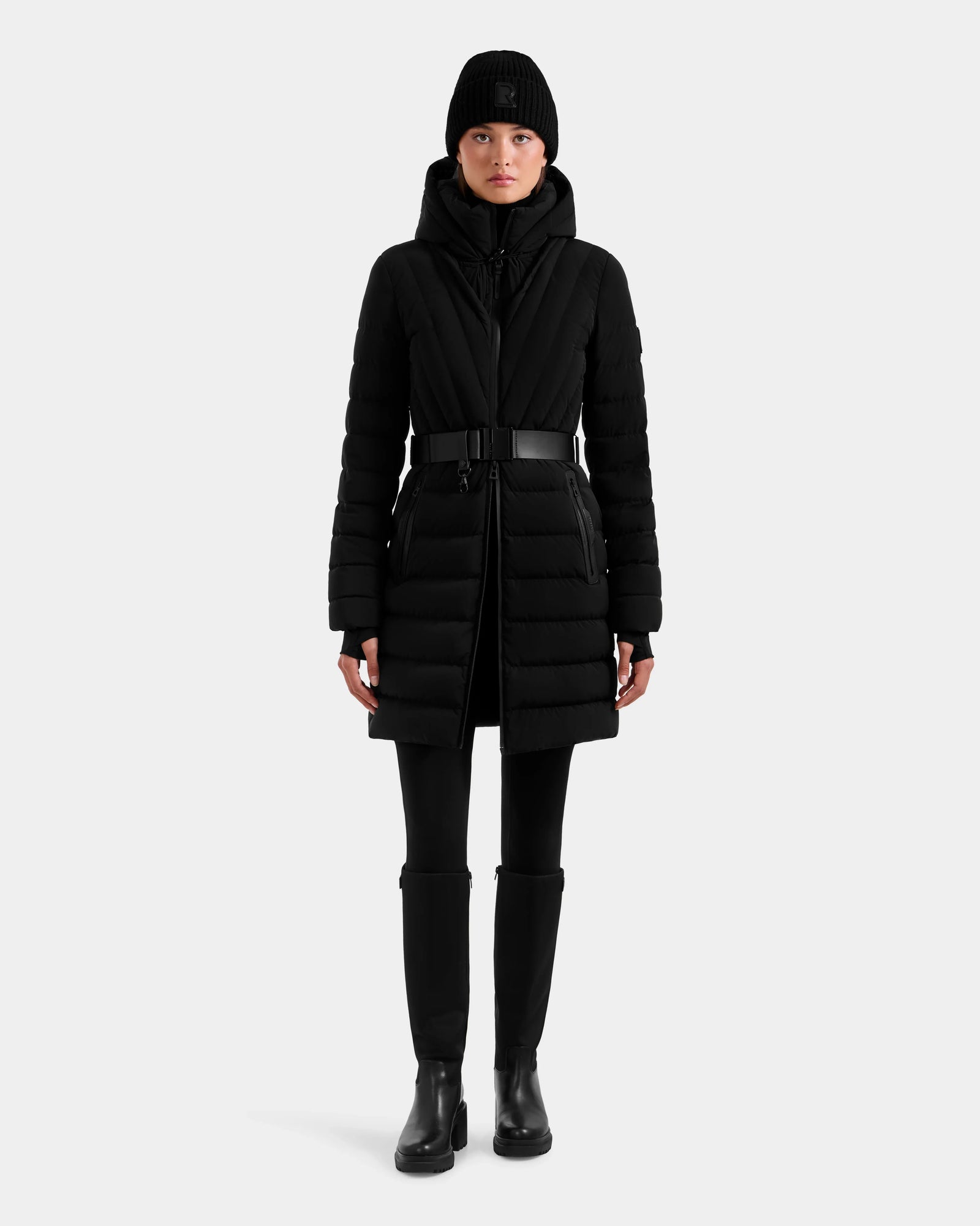 A person wearing the Rudsak ALISA M - L125586 Black long quilted down parka with a vegan leather belt, black pants, boots, and a beanie stands facing forward against a plain light gray background.