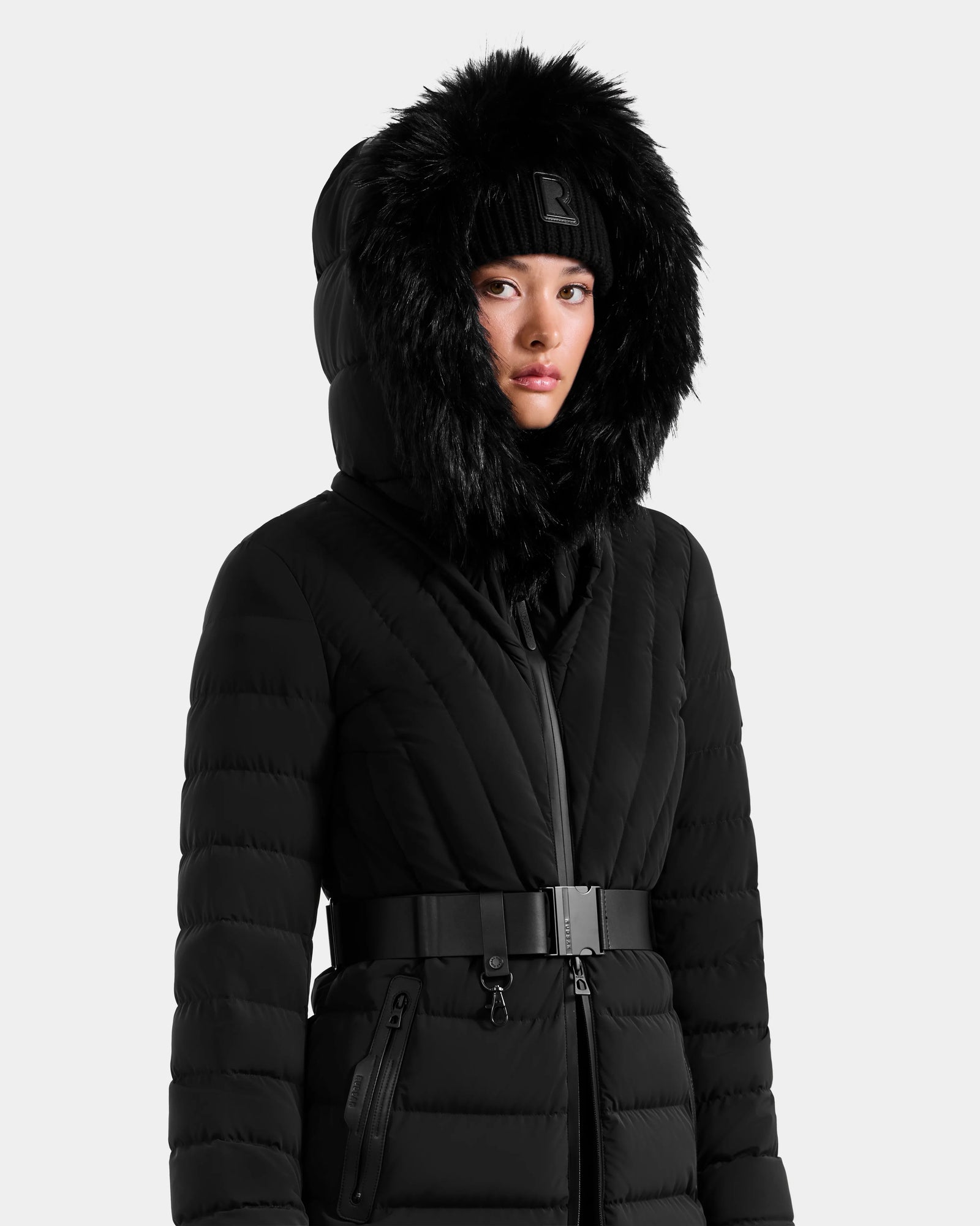 A woman wears the Rudsak ALISA M - L125586 Black quilted down parka with a black faux fur-trimmed hood, paired with a matching black beanie and wide belt, standing against a plain light background.