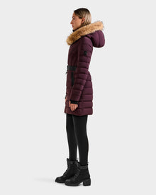 A woman stands in profile against a plain light background, wearing the Rudsak ALISA M - L125586 Bordeaux long dark purple quilted down parka with a faux fur hood, paired with black leggings and black lace-up boots.