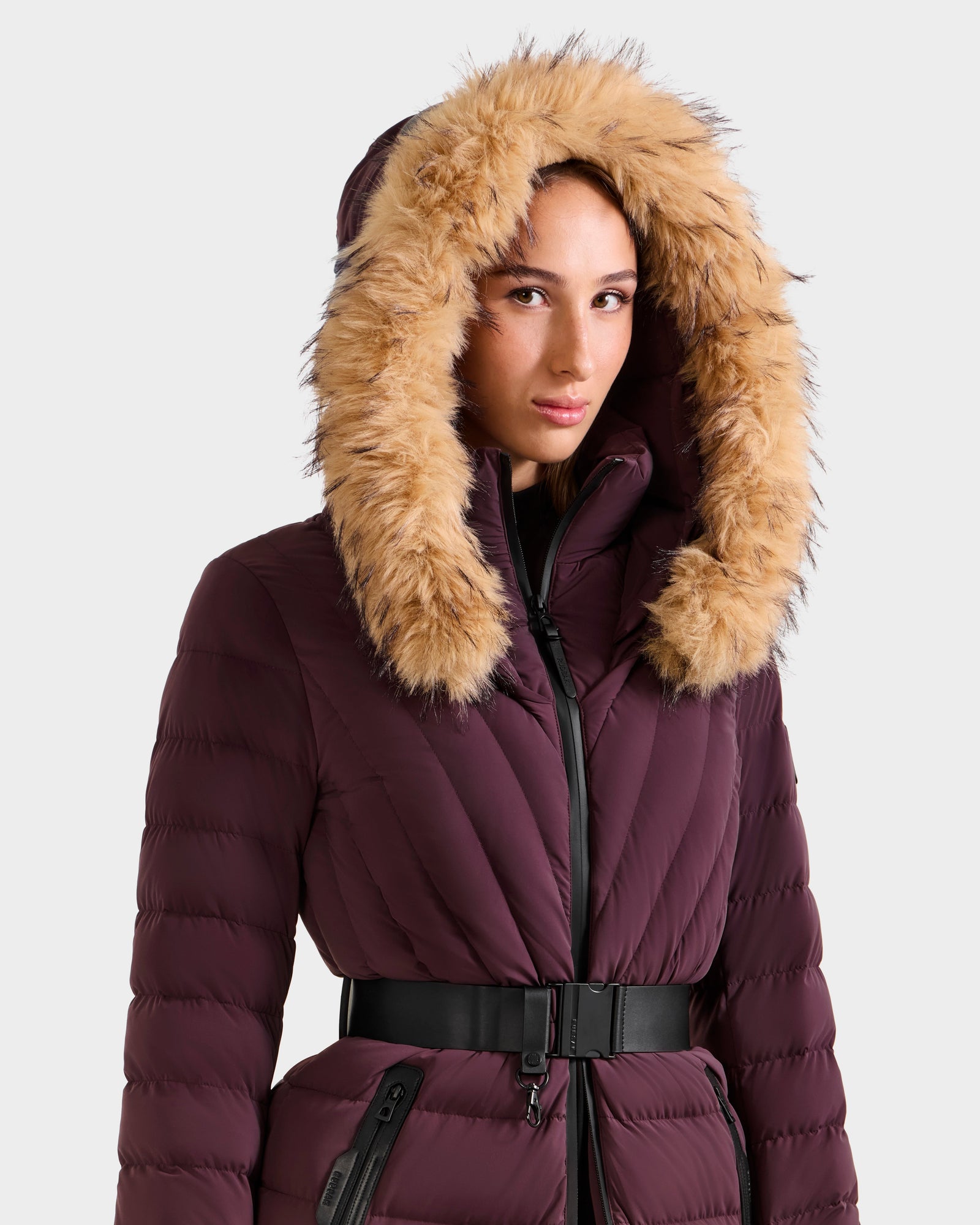 A woman wears the Rudsak ALISA M - L125586 Bordeaux, a maroon quilted down parka with a faux fur hood and black vegan leather belt, standing against a plain light background.