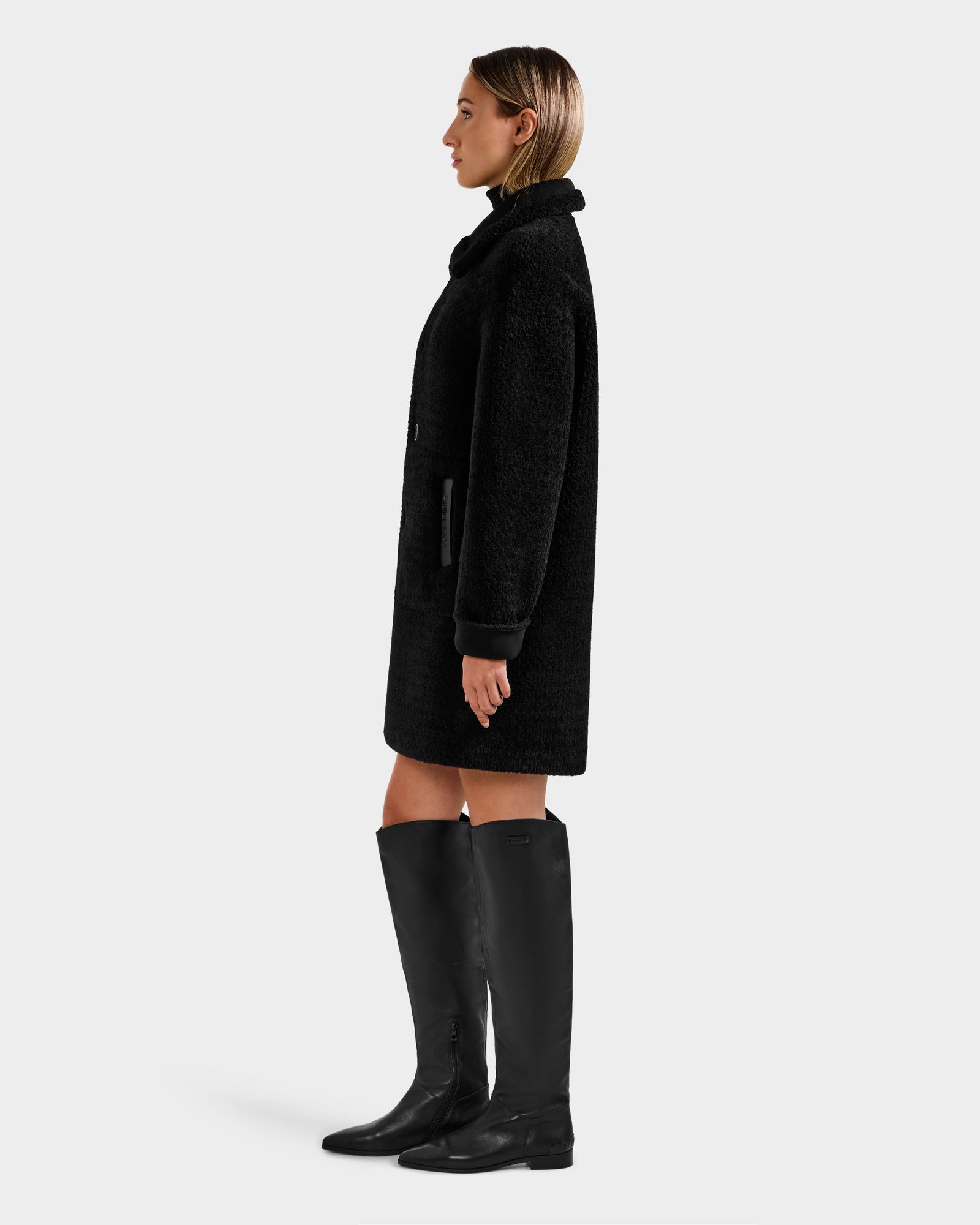 A woman in profile against a plain background wears the Rudsak ANGILICA - L125600 Black faux shearling knee-length coat with a textured finish and black knee-high boots. Her straight hair is parted in the middle.