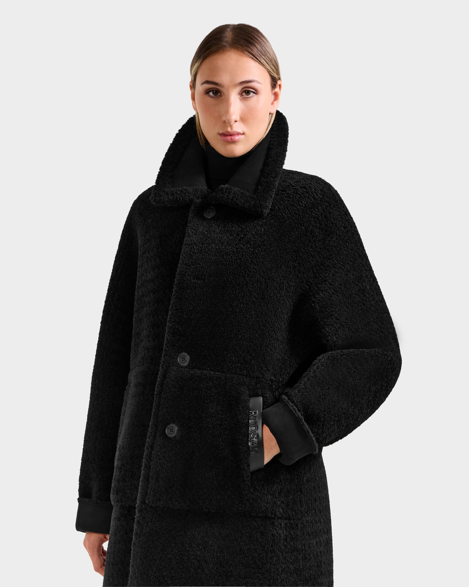 A woman with light brown hair models the ANGILICA - L125600 Black faux shearling coat by Rudsak, featuring large buttons and a high collar, against a plain light gray background.