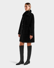 A woman models the Rudsak ANGILICA - L125600 Black faux suede coat with large front pockets and a high collar, paired with black knee-high boots. She has straight, shoulder-length hair and stands against a plain light gray background.