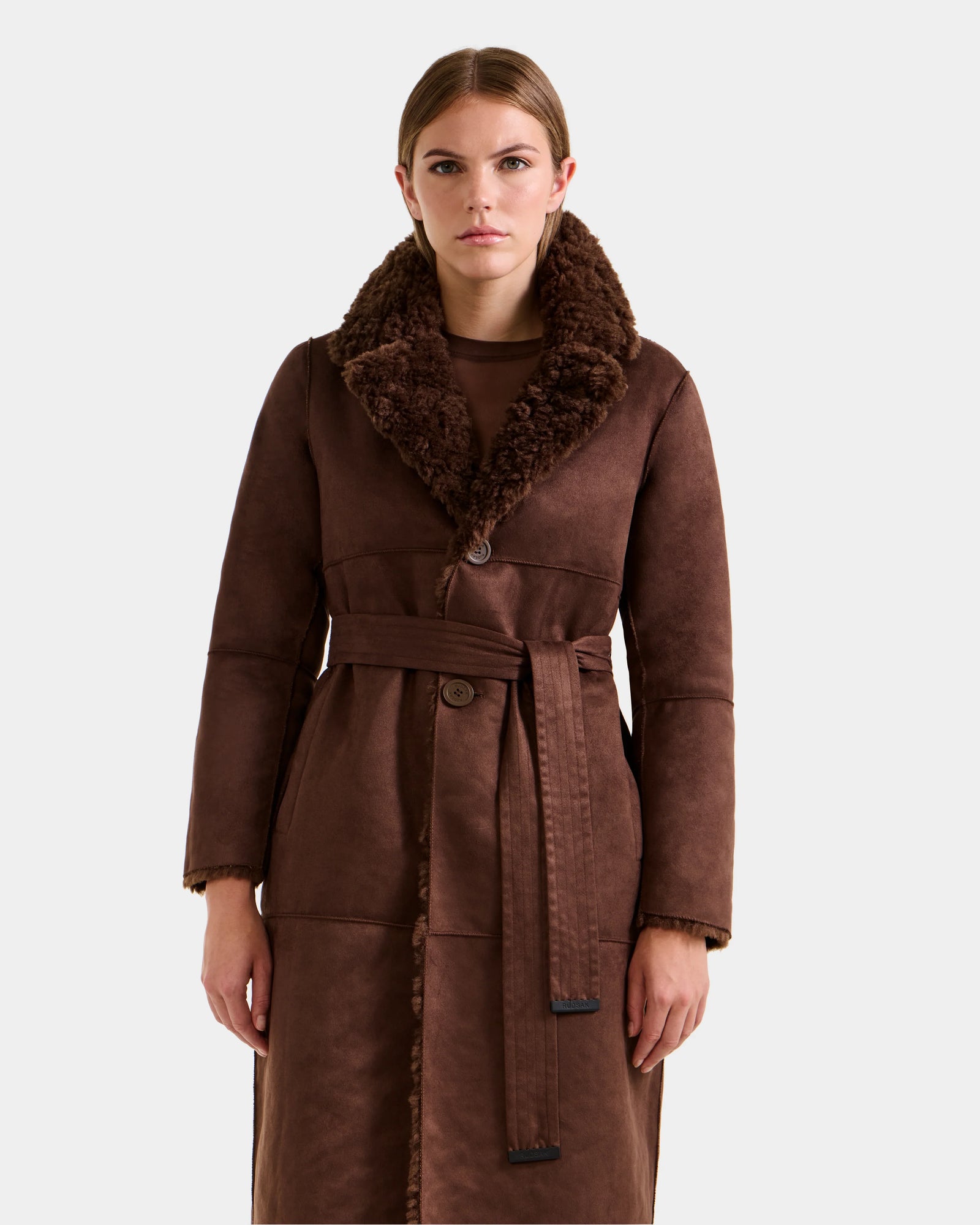 A woman wears the Rudsak ANNABELLE FSU - L125512 Espresso, a long brown faux suede coat with a textured sherpa teddy collar and matching belt, standing against a plain light grey background.